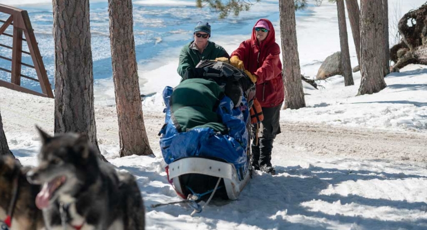 two people stand behind a sled in a snowy landscape. There are dogs in the foreground. 